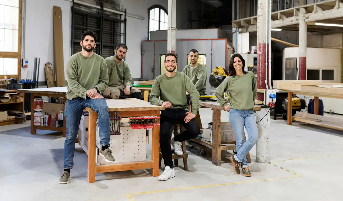 Fünf Mitarbeitende eines Handwerksbetriebs in grünen Sweatshirts posieren lächelnd in einer hellen Tischlerei-Werkstatt – Personalvermittlung für das Handwerk mit Matchmeister